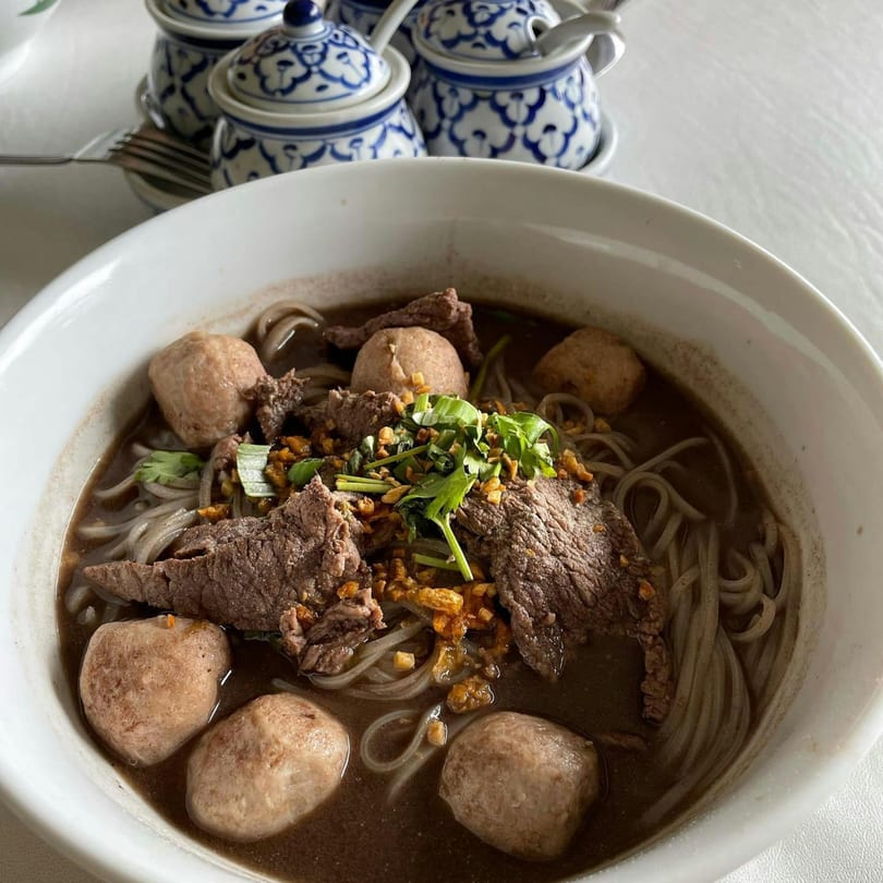 A bowl of Kook Kai's Thai Noodles with beef, meatballs, and herbs on a white table.